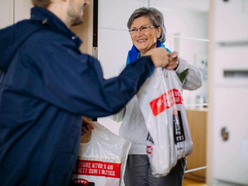 Bestemor mottar varer fra en person i en butikk, med handleposer i hendene.