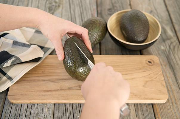 Person skreller en avocado på et skjærebrett med en kniv, mens en annen avocado ligger i en skål i bakgrunnen.
