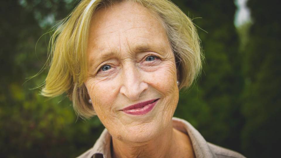 En person med blond hår står utendørs med grønt løv i bakgrunnen.