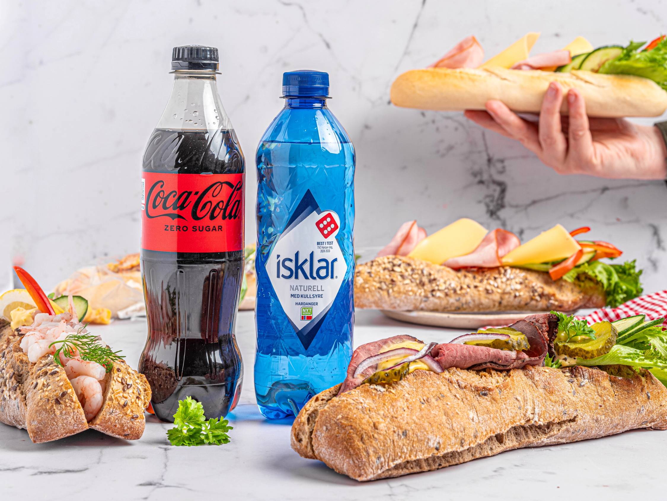 To flasker, en Coca-Cola og en isklar vannflaske, står foran et utvalg av baguetter med pålegg på et bord.