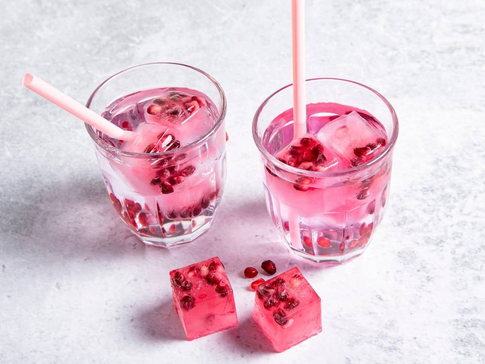 To glass med isbiter og granateplekjerner, med sugerør i hver, plassert på en lys overflate.