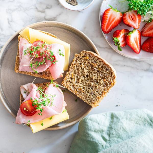 To glutenfrie matpakkebrød med pålegg, en med skinke og ost og en med skinke, ost og tomat, på en tallerken med jordbær og en glass melk ved siden av.