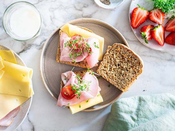 To glutenfrie matpakkebrød med pålegg, en med skinke og ost og en med skinke, ost og tomat, på en tallerken med jordbær og en glass melk ved siden av.