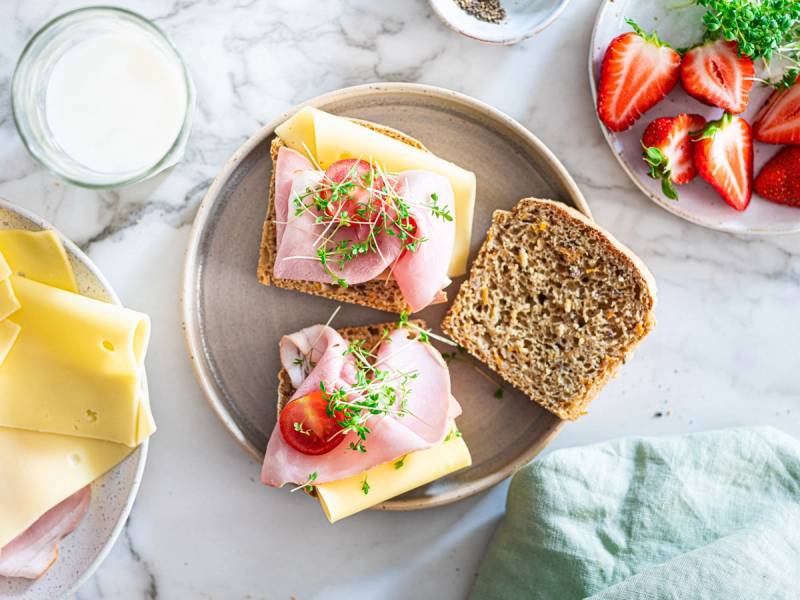 To glutenfrie matpakkebrød med pålegg, en med skinke og ost og en med skinke, ost og tomat, på en tallerken med jordbær og en glass melk ved siden av.
