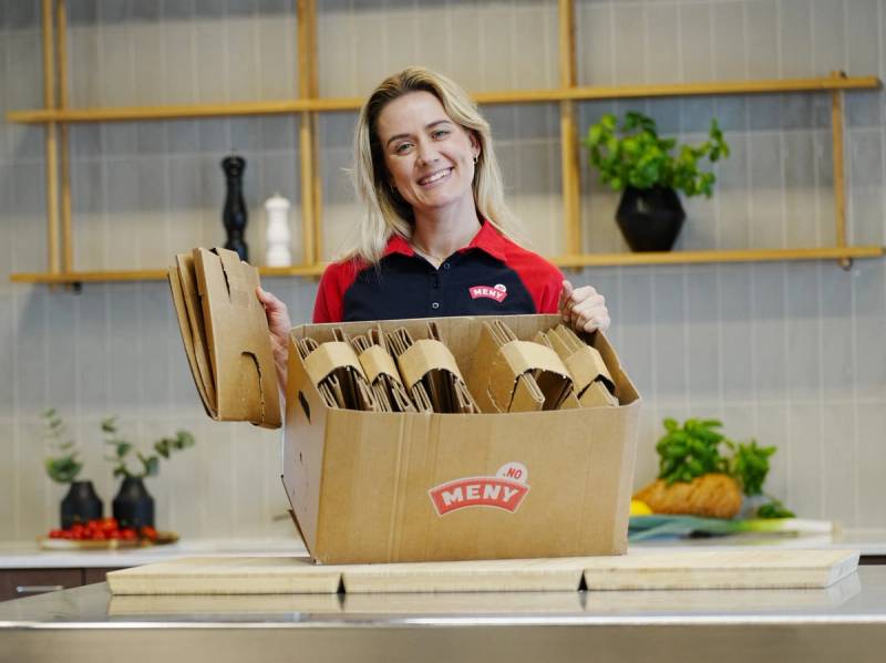 En person med ansiktet dekket står bak en åpen eske med brød på et kjøkken, med hyller og planter i bakgrunnen.