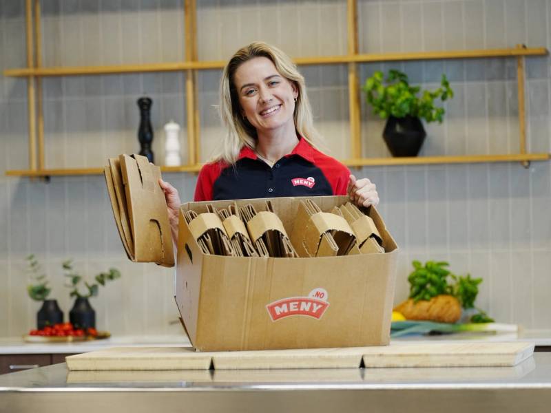 En person med ansiktet dekket står bak en åpen eske med brød på et kjøkken, med hyller og planter i bakgrunnen.