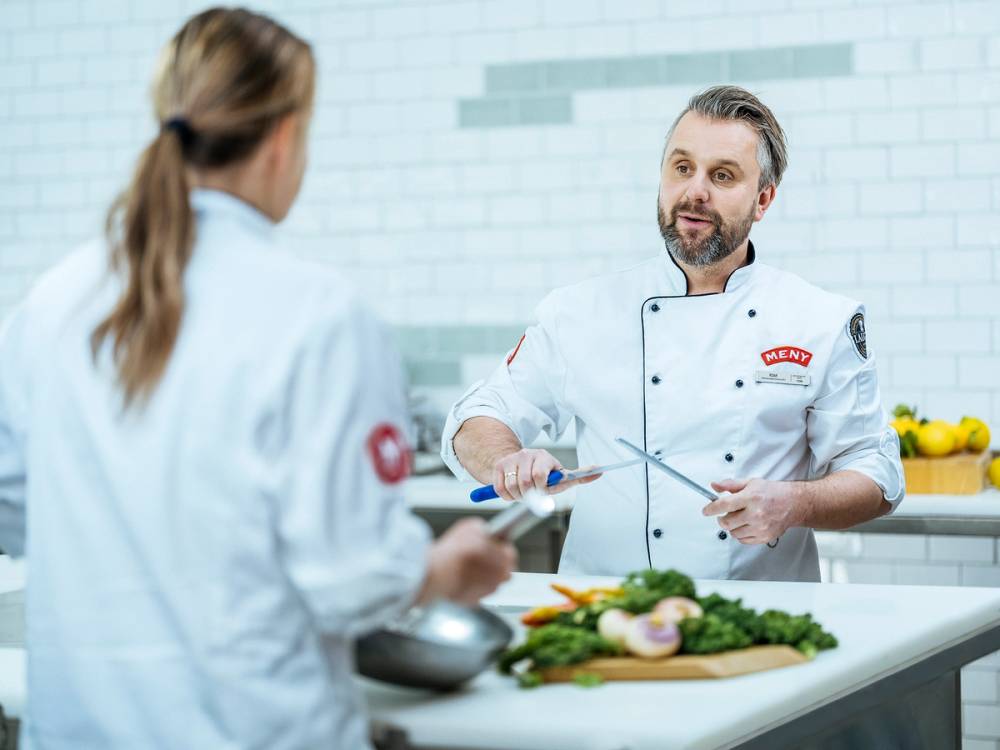 To personer i kokkeklær som snakker på kjøkken med grønnsaker og kjøkkenutstyr foran seg.