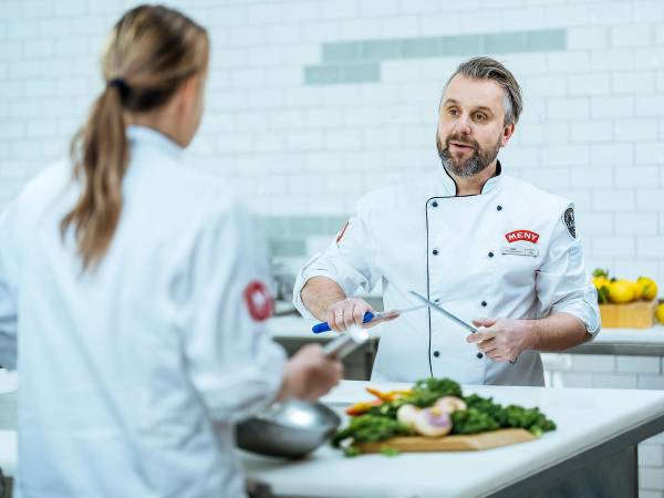 To personer i kokkeklær som snakker på kjøkken med grønnsaker og kjøkkenutstyr foran seg.