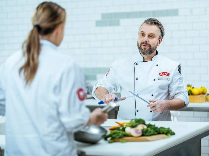 To personer i kokkeklær som snakker på kjøkken med grønnsaker og kjøkkenutstyr foran seg.