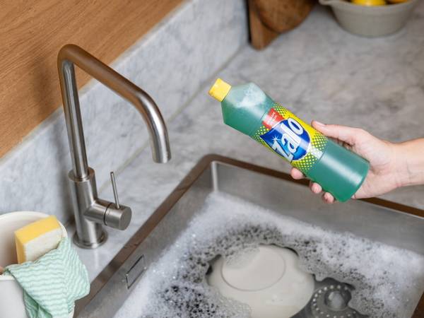 En person holder en flaske Zalo opp over en oppvaskkum med oppvaskmiddel og skummer, klar til å helle inn i vasken.