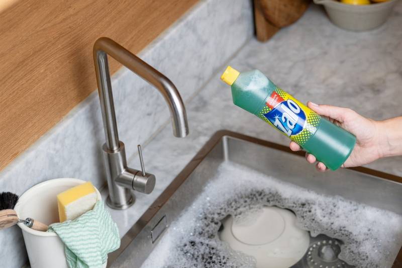 En person holder en flaske Zalo opp over en oppvaskkum med oppvaskmiddel og skummer, klar til å helle inn i vasken.