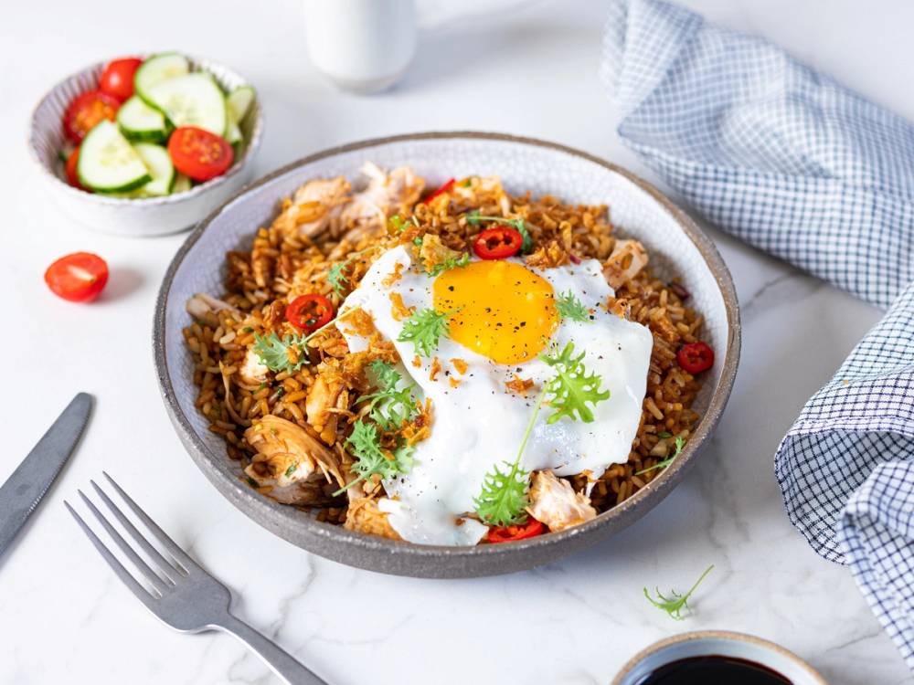Bolle med nasi goreng toppet med et speilegg, chili, og grønnsaker, omgitt av en liten salat og en kopp kaffe på et hvitt bord.