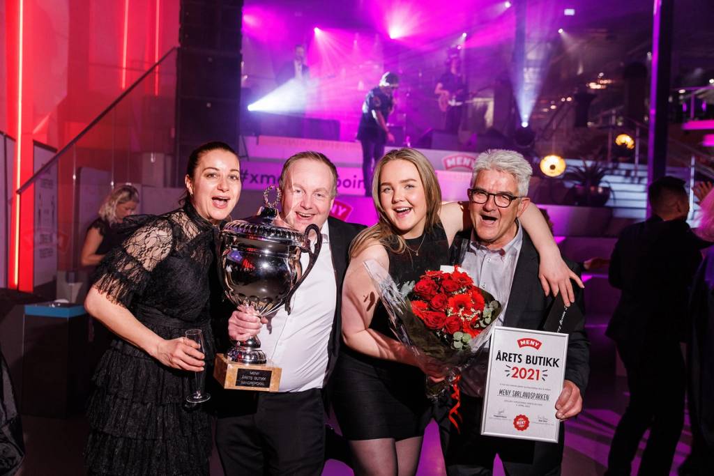 Fire personer på scenen som feirer med et trofé og blomster, med en fargerik lyssetting i bakgrunnen.