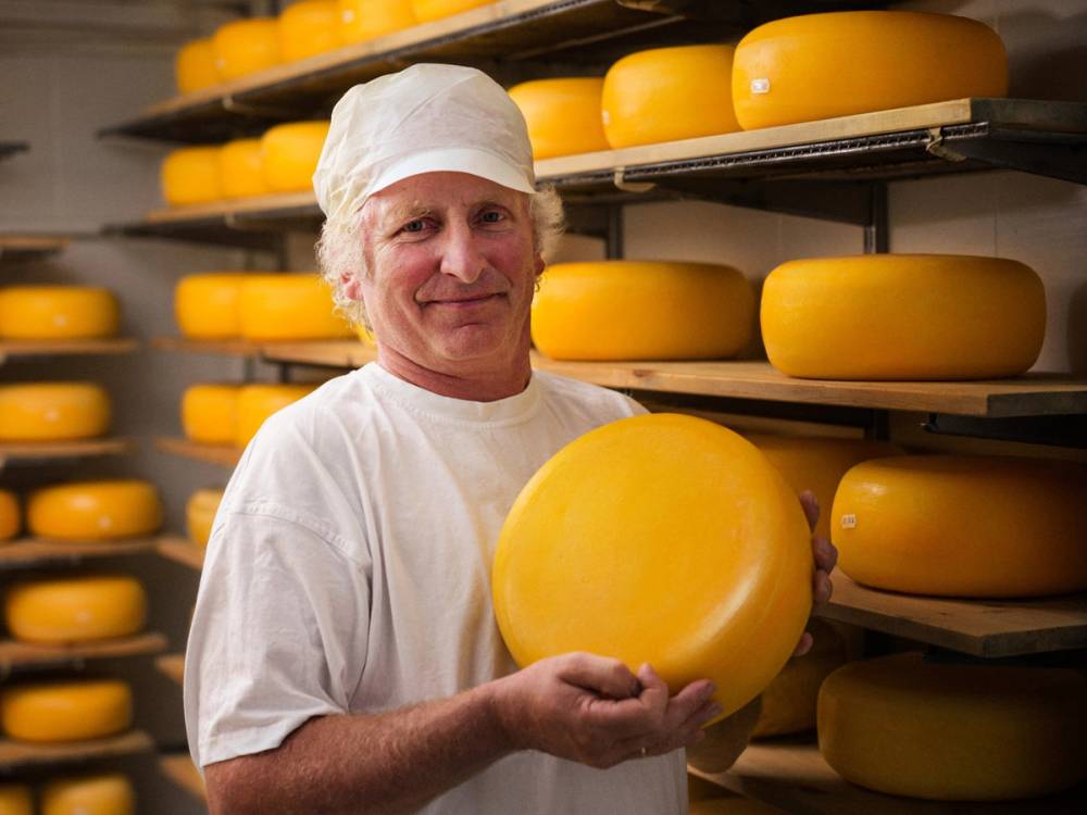 Person i hvit kjeledress som holder et stort gult ostestykke i en ostebutikk eller ostelager med hyller fulle av ost.