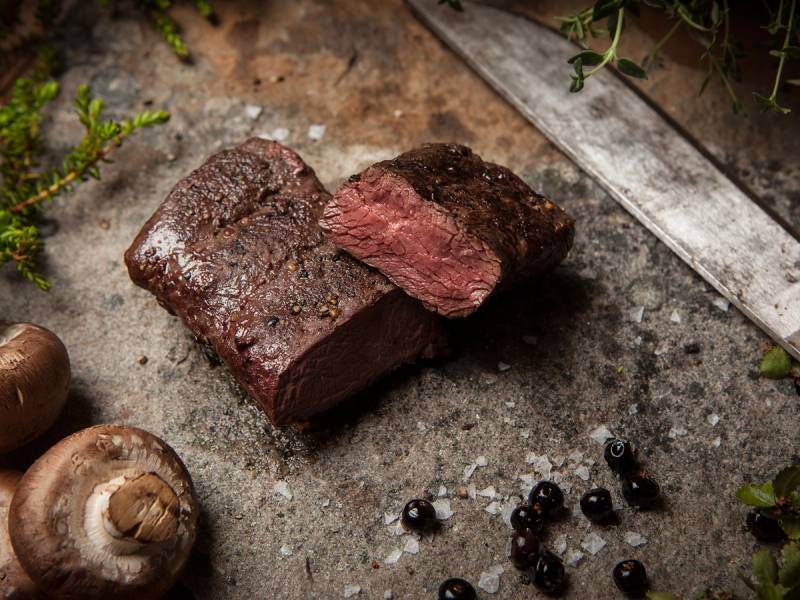 Kjøttstykker som er stekt på naturlige premisser, med sopp og svarte bær rundt på en stekeplate.