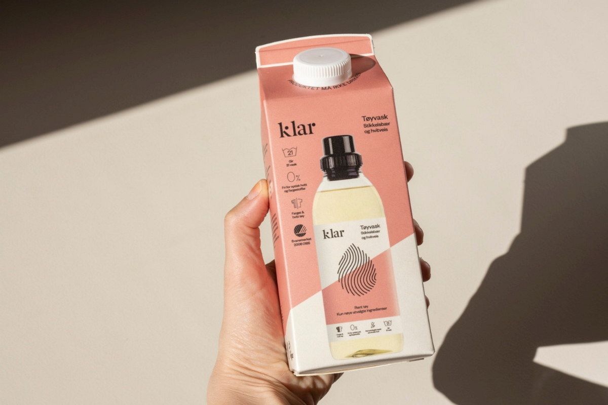 En hånd holder en kartong med drikke, der står 'Klar' på pakken, og det er en liten flaske inni kartongen.