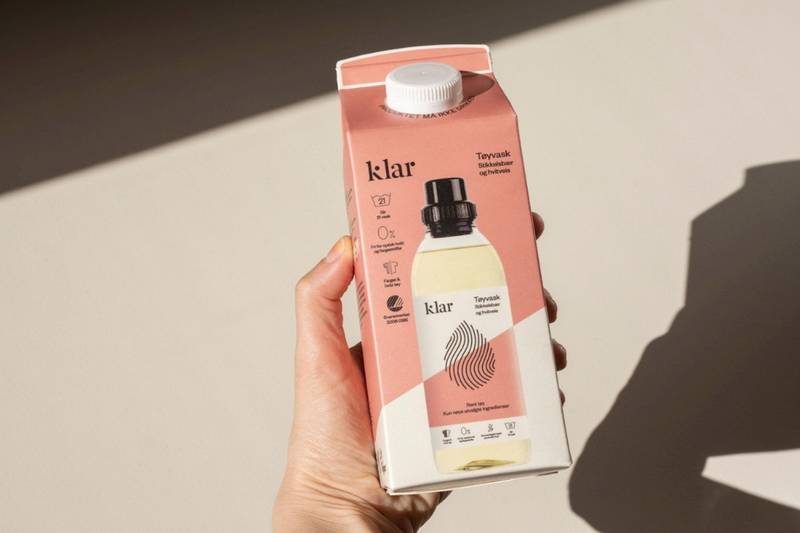 En hånd holder en kartong med drikke, der står 'Klar' på pakken, og det er en liten flaske inni kartongen.