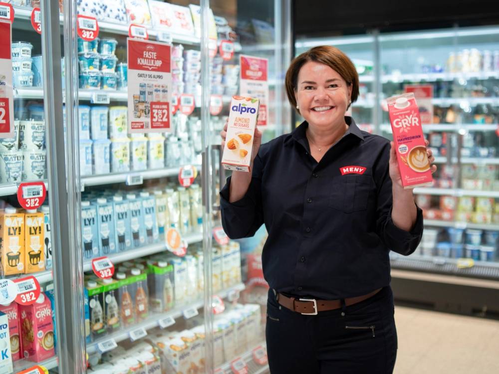 En person i butikk holder opp to ulike melkeprodukter, en i hver hånd, foran kjøledisk med meieriprodukter.