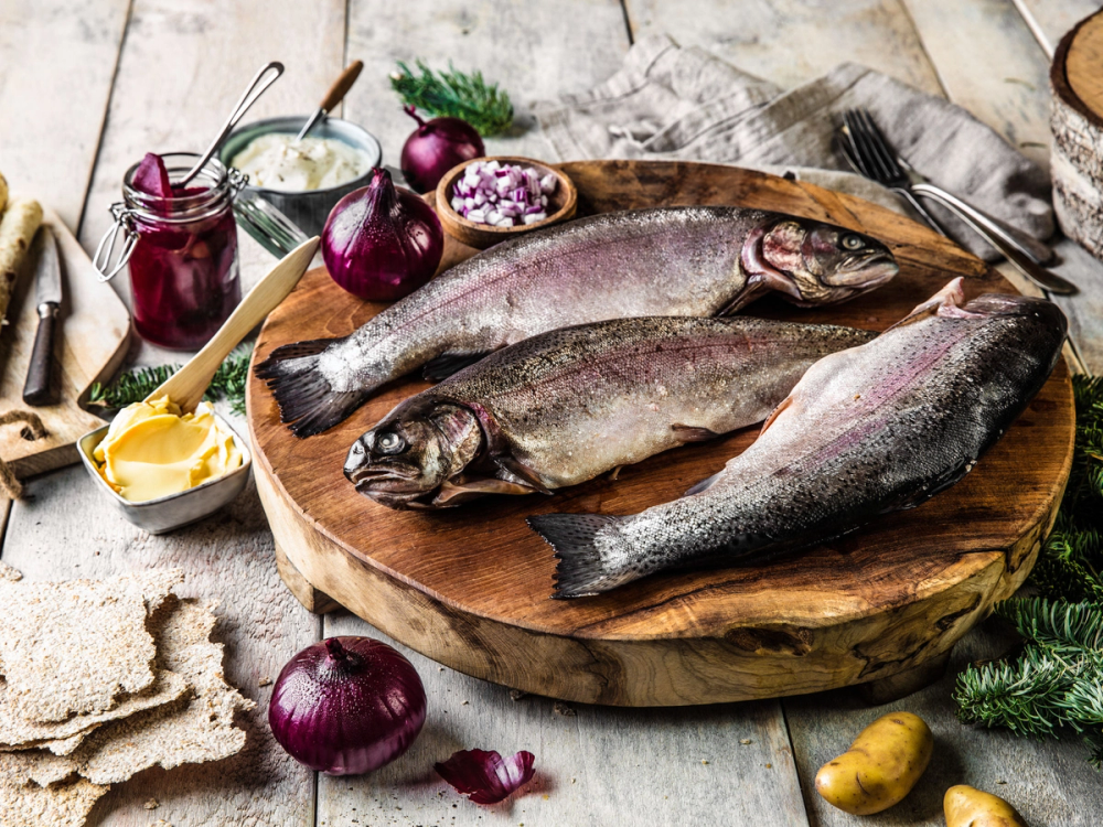 Tre rakfisk ligger på et rundt trebrett med tilbehør som rødløk, smør og sylteagurk rundt, klar til servering.