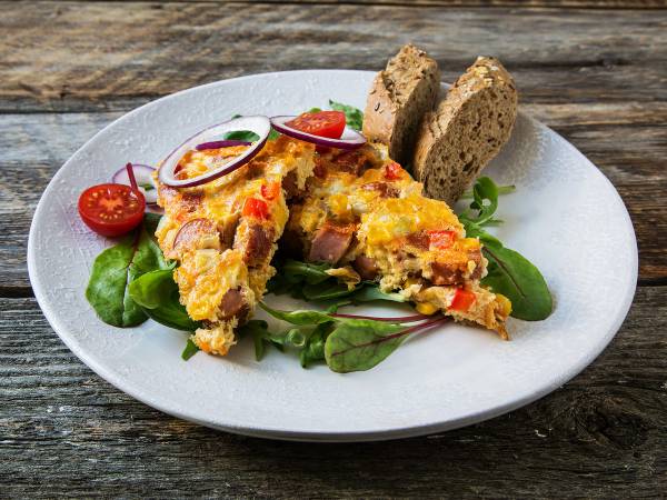 En porsjon pølseform med ost, tomat, løk og salat på en hvit tallerken, med to skiver brød ved siden av, på et trebord.