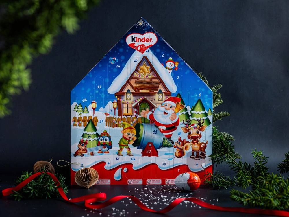 En julekalender med julemotiver og snø, plassert mot en mørk bakgrunn med grønne grener rundt.