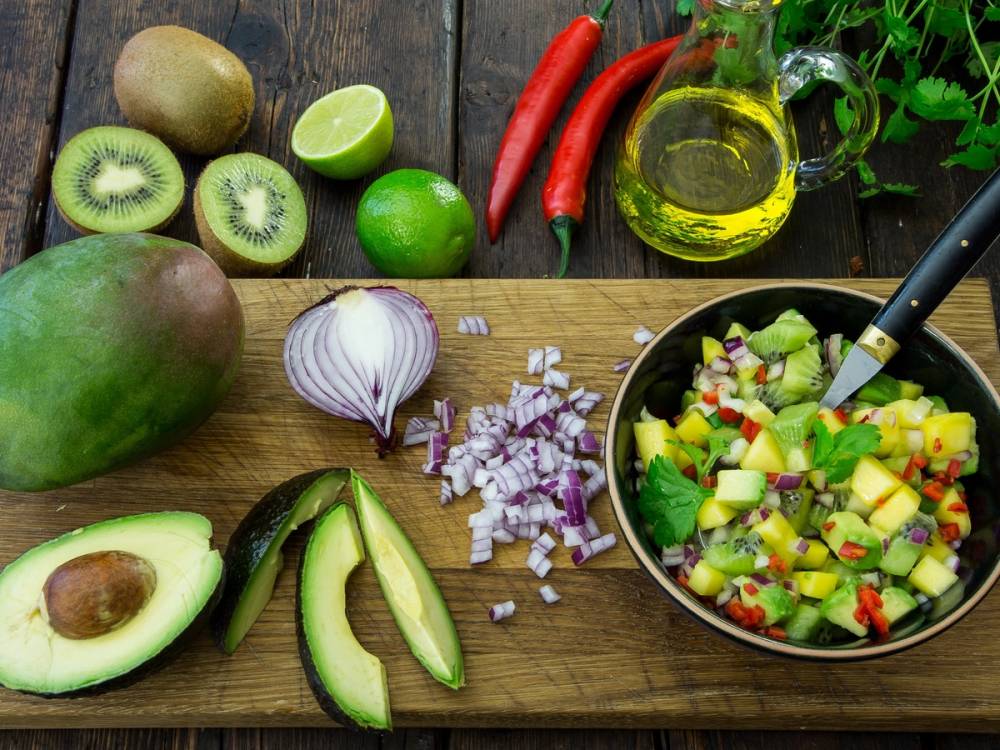 Skjærebrett med oppkuttet avokado, løk og en bolle med salat med frukt og grønnsaker, omgitt av hele kiwi, lime, chili, olje og urter.