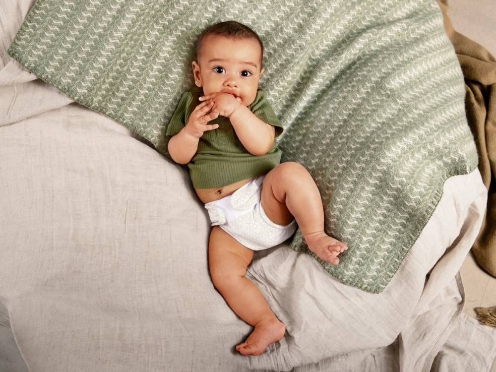 En baby ligger på en seng med grønne og beige puter, iført en hvit bleie og en grønn t-skjorte, og holder en finger i munnen.