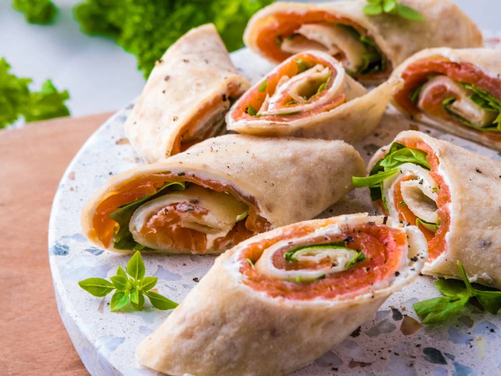 Lefseruller med røkelaks, kremost og grønnsaker på en hvit tallerken, pyntet med urter.