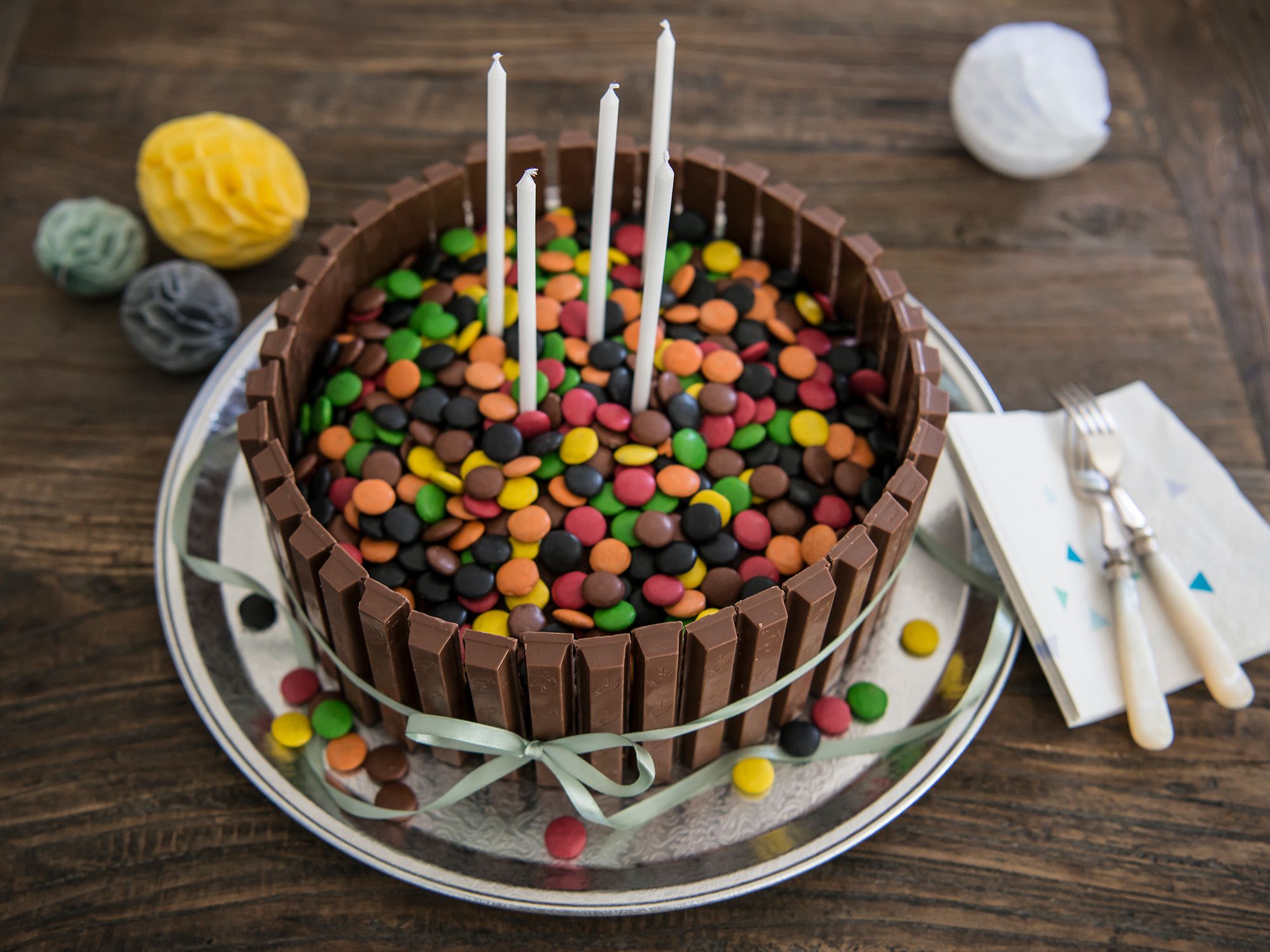 En kake dekorert med sjokoladeplater og fargerike smågodt, med tre lys på toppen, plassert på et fat med pynt og bestikk rundt.