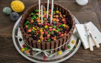 En kake dekorert med sjokoladeplater og fargerike smågodt, med tre lys på toppen, plassert på et fat med pynt og bestikk rundt.