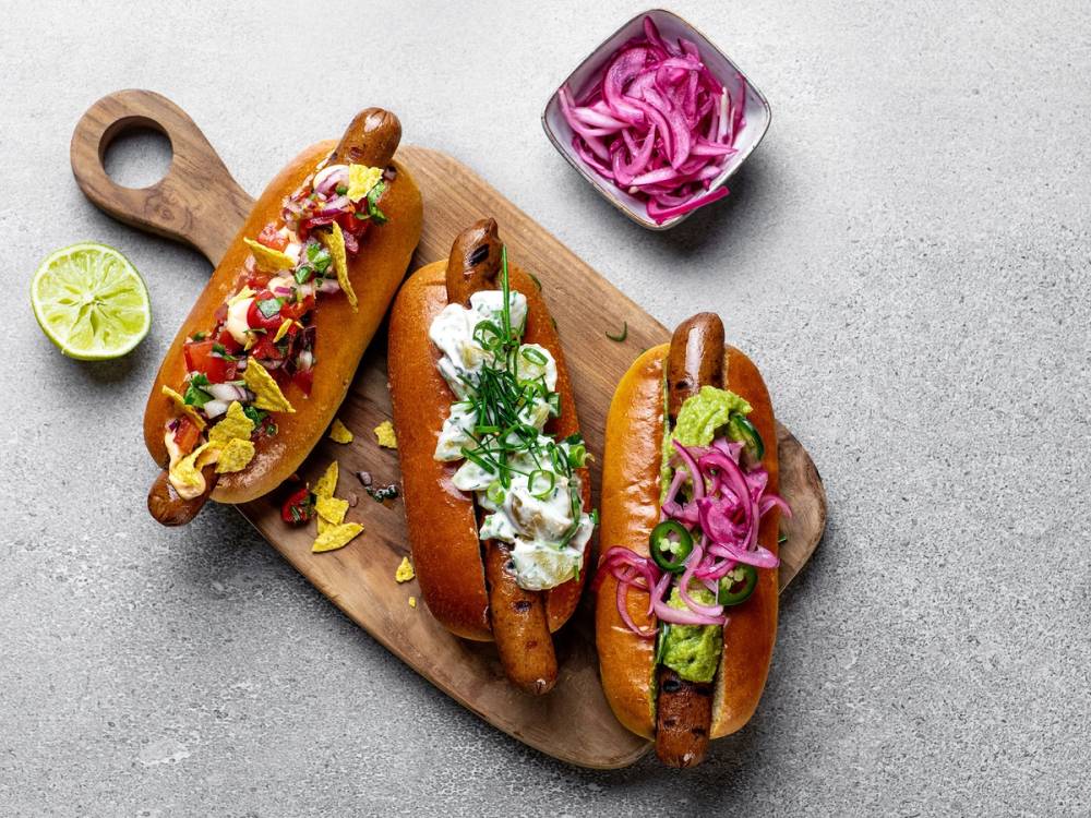 Tre pølser i brød med ulike tilbehør på en treplate, med en liten skål med syltet rødløk ved siden av.