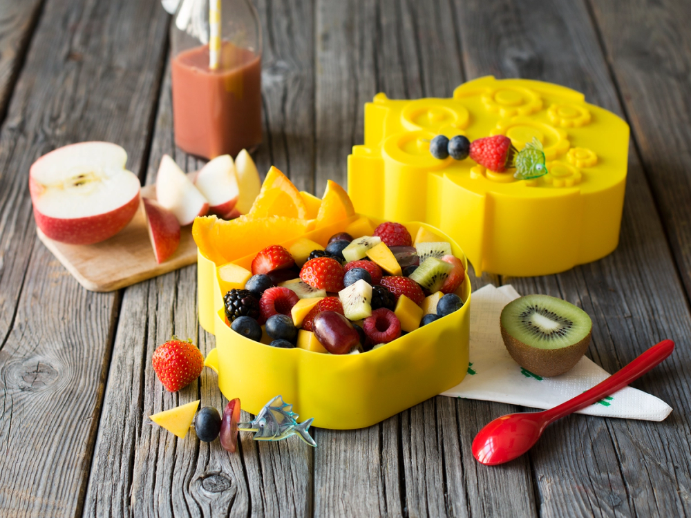Fargerik fruktsalat i en gul form med fersk frukt og bær, omgitt av skiver av kiwi, eple og en liten skål med juice på et trebord.