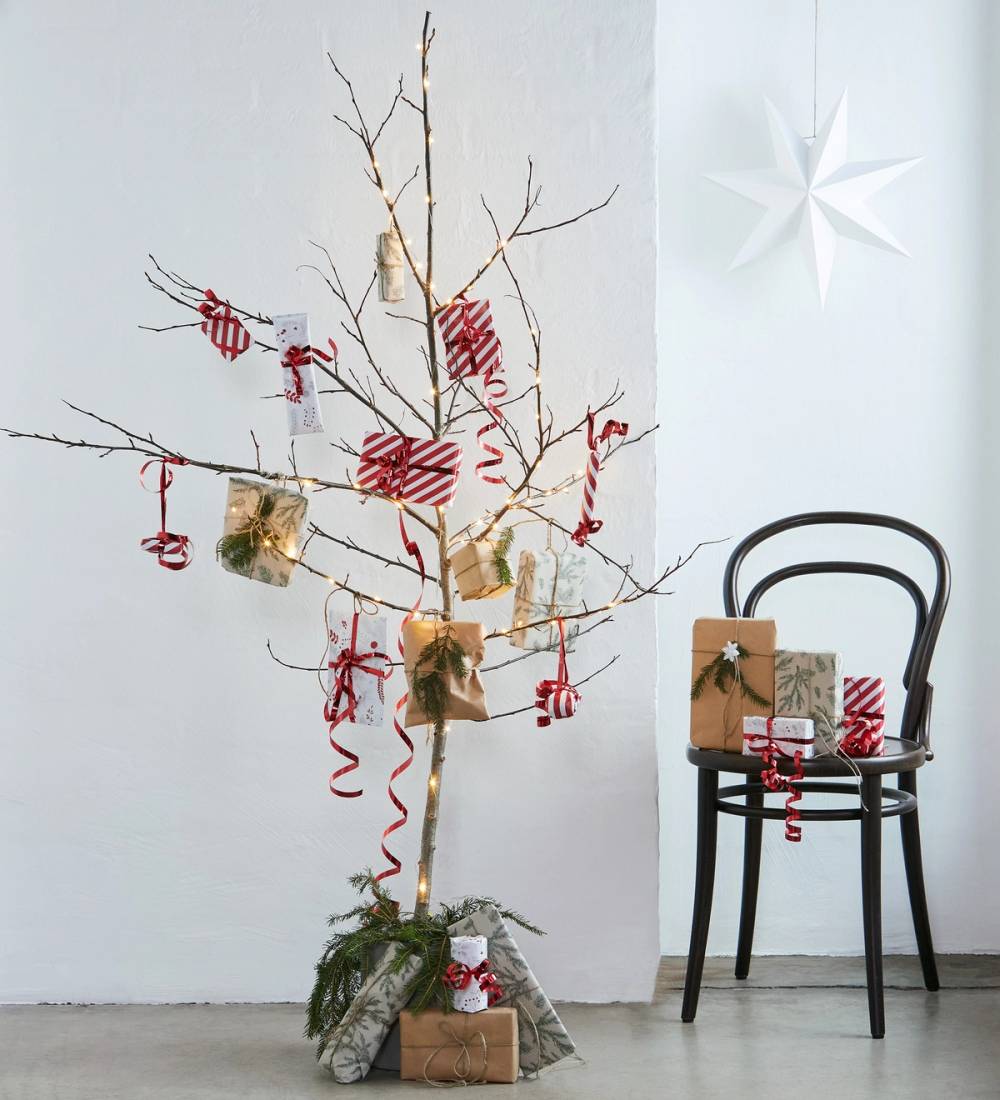 Et dekorert juletre uten blader med gaver under, plassert ved en svart stol med julepynt og pakker på, i et lyst rom med hvit vegg og en stjerne på veggen.