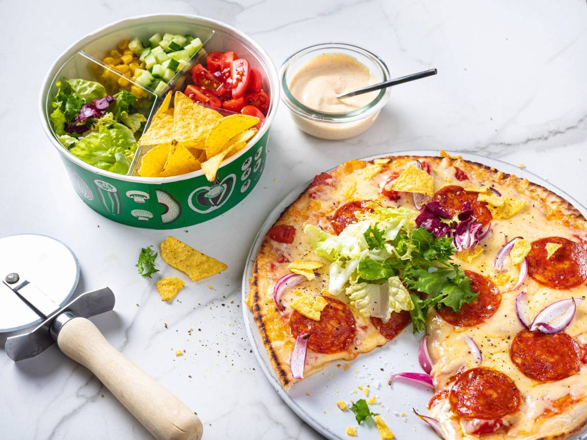 En pizza med pepperoni, løk og salat på en tallerken, med en salatbar med grønnsaker og en dressing ved siden av.