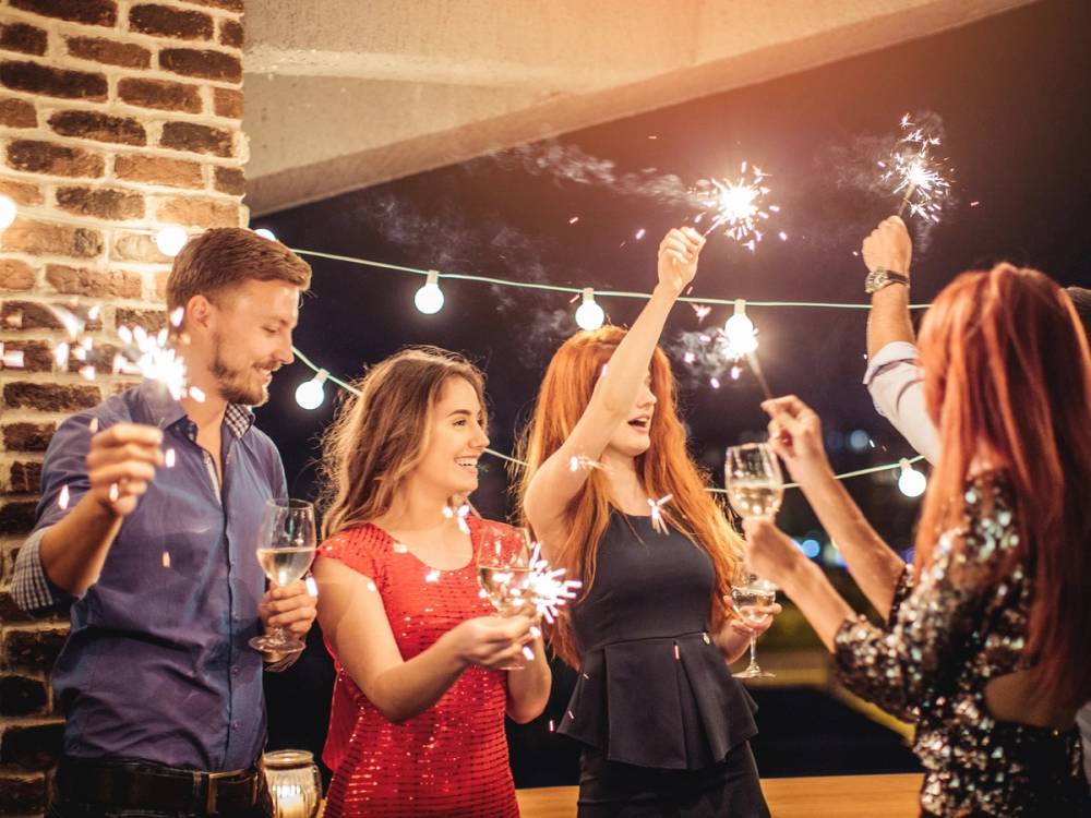 En gruppe mennesker feirer med fyrverkeri og drikker vin innendørs, pyntet med lyslenker og en murvegg i bakgrunnen.
