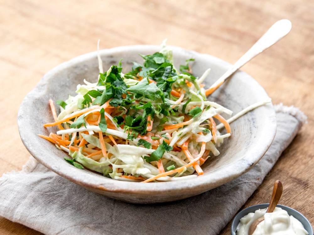 En bolle med coleslaw bestående av revet kål og gulrøtter, pyntet med frisk grønnkål, på et trebord med en skje og en liten skål med saus ved siden av.