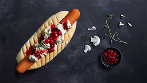 En hotdog med pølse, ketchup, sennep og løk på en lang bolle, plassert på en svart overflate med noen sopp, urter og en liten skål med rød saus ved siden av.