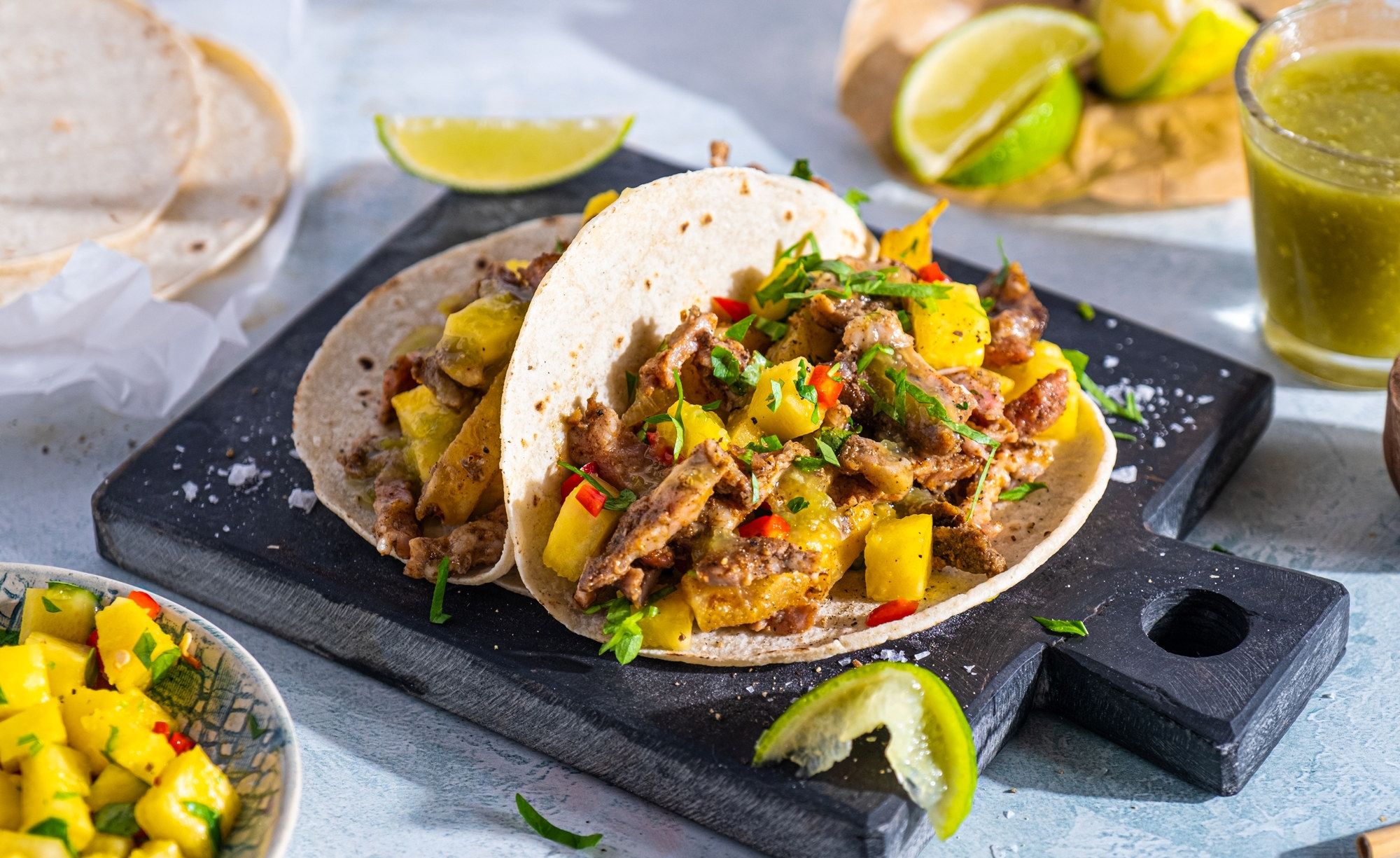 To soft tacos fylt med svinekjøtt, grønnsaker og koriander, servert på en svart skjærefjøl med limebåter og en grønn saus i bakgrunnen.
