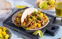 To soft tacos fylt med svinekjøtt, grønnsaker og koriander, servert på en svart skjærefjøl med limebåter og en grønn saus i bakgrunnen.