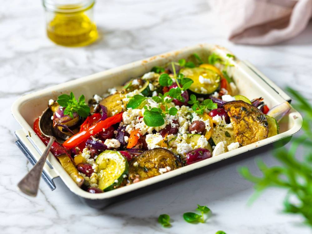 En ovnsbakt grønnsakssalat med aubergine, paprika, squash, oliven og fetaost i en hvit ildfast form.