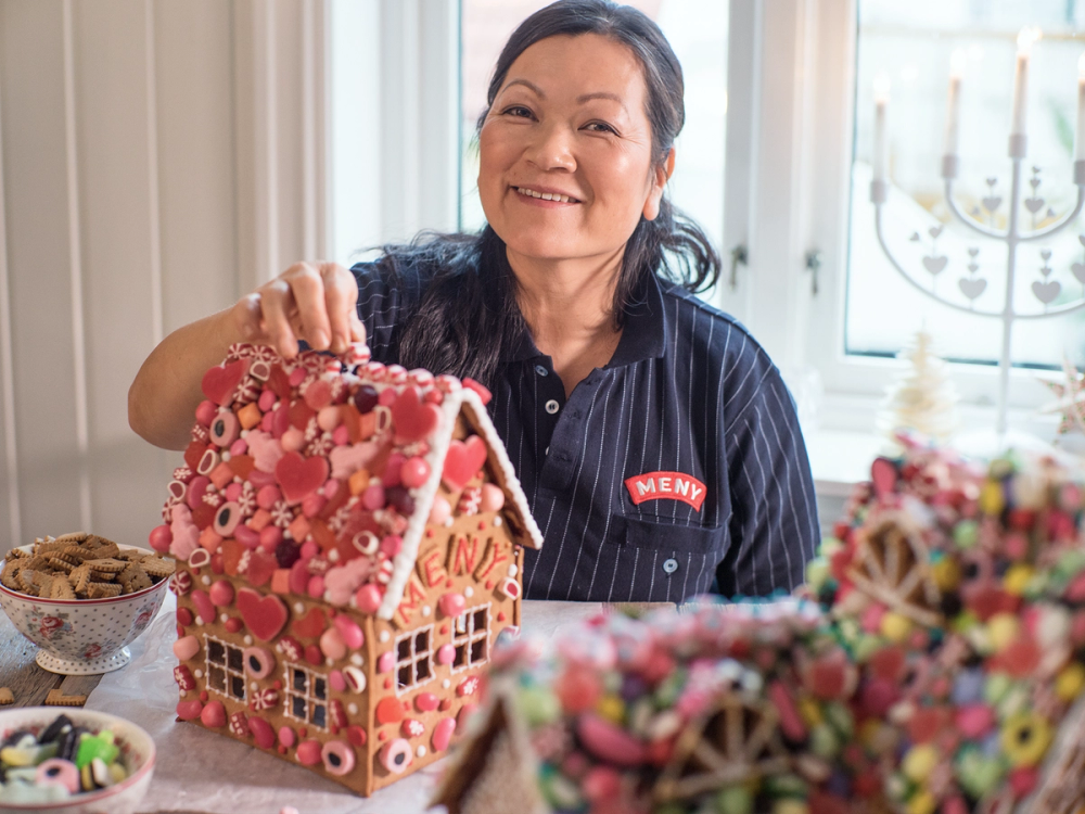 En person som lager eller viser et pepperkakehus dekorert med rosa og hvite blomster og andre pynt, i et lyst rom med store vinduer.