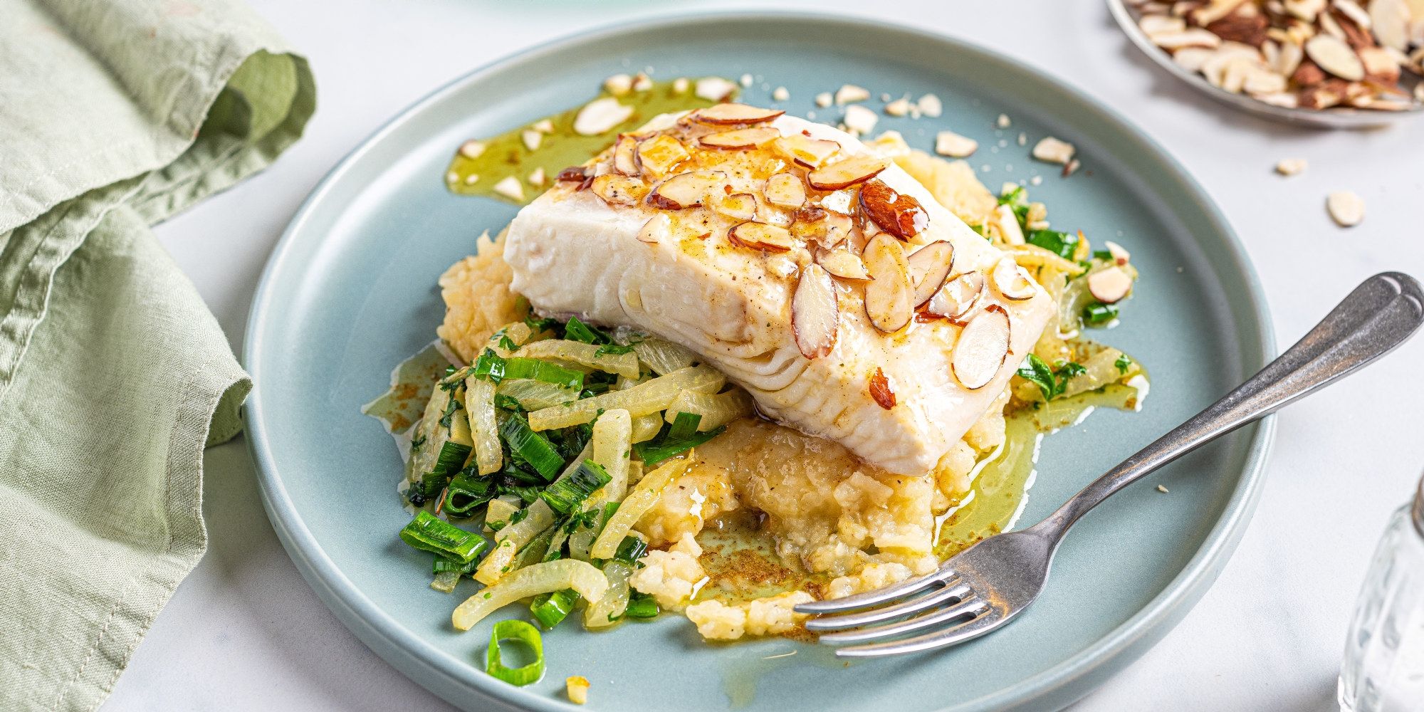 En tallerken med stekt kveitefilet toppet med skiver av mandler, servert med potetmos og grønnsaker, på et lyst bord.