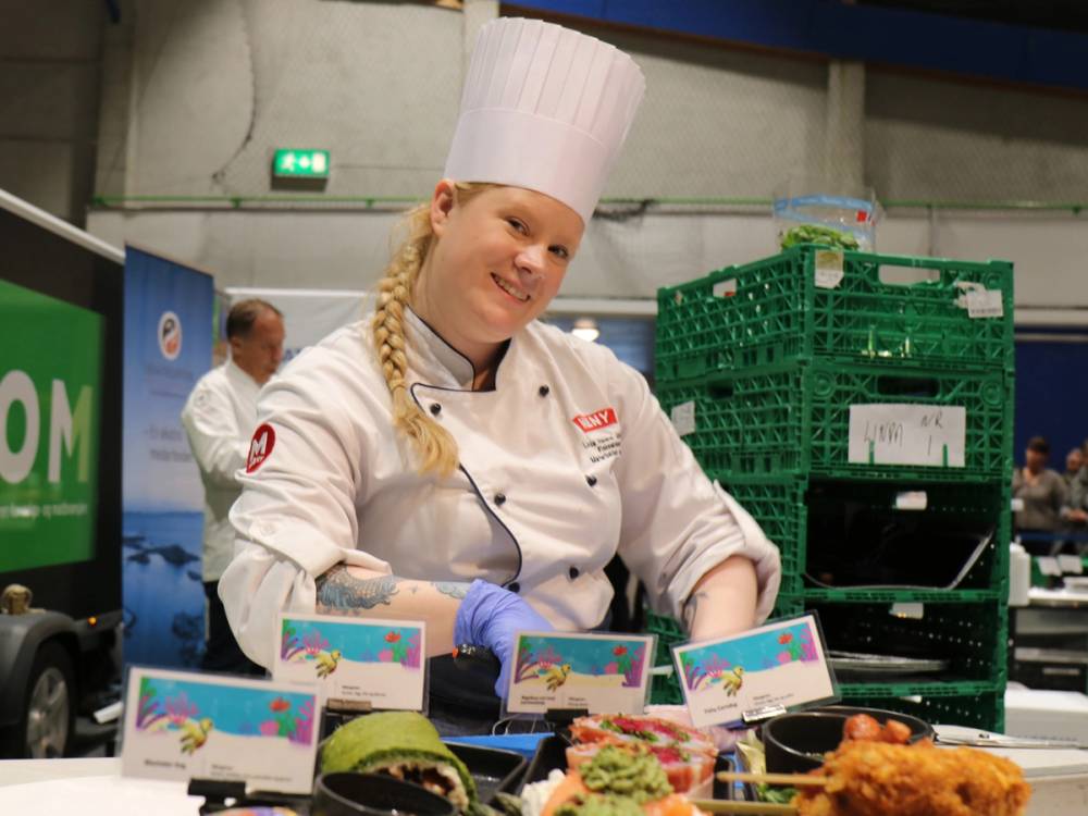 En person i kokkeforkle og hvit kokkejakke står bak en matbod med ulike retter og kort, på en messe eller utstilling, med grønne plastkasser i bakgrunnen.