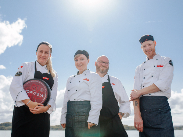 Fire personer i kokkeklær står ute under en klar himmel, med en meny for oste-VM i hånden til en av dem.
