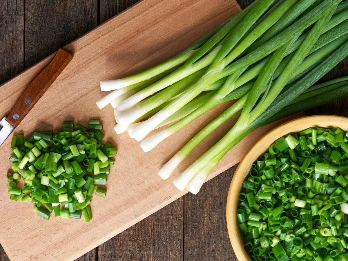 Skjæreflate med hakket vårløk og hele vårløk på et skjærebrett og en skål med hakket vårløk, på et trebord.