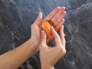 To hender som legger en skive med laks på en liten kloss av ris for å lage nigiri, mot en mørk steinbakgrunn.