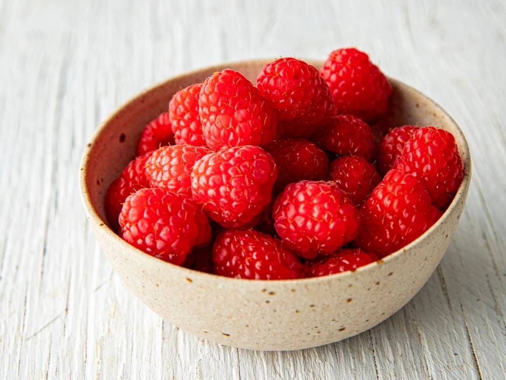 En liten keramisk bolle fylt med friske, røde bringebær på et lyst treverk.