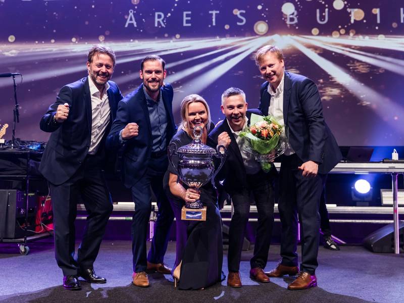 Fem personer i dress står på en scene med en stor skjerm bak dem, og en av dem holder en pokal og blomsterbukett, sannsynligvis etter en premieutdeling.