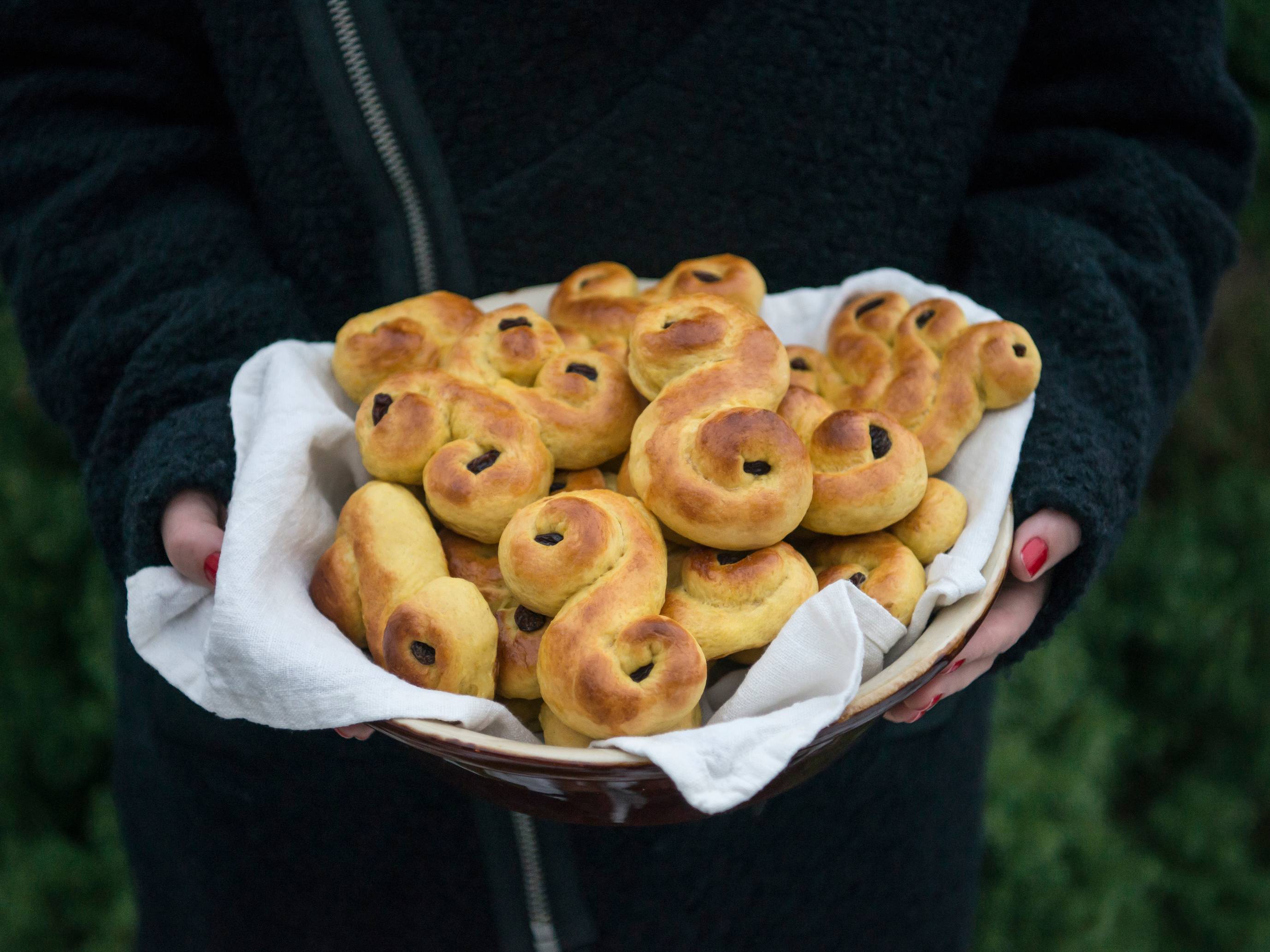 Person holder en tallerken med nybakte lussekatter på, omgitt av grønt gress i bakgrunnen.