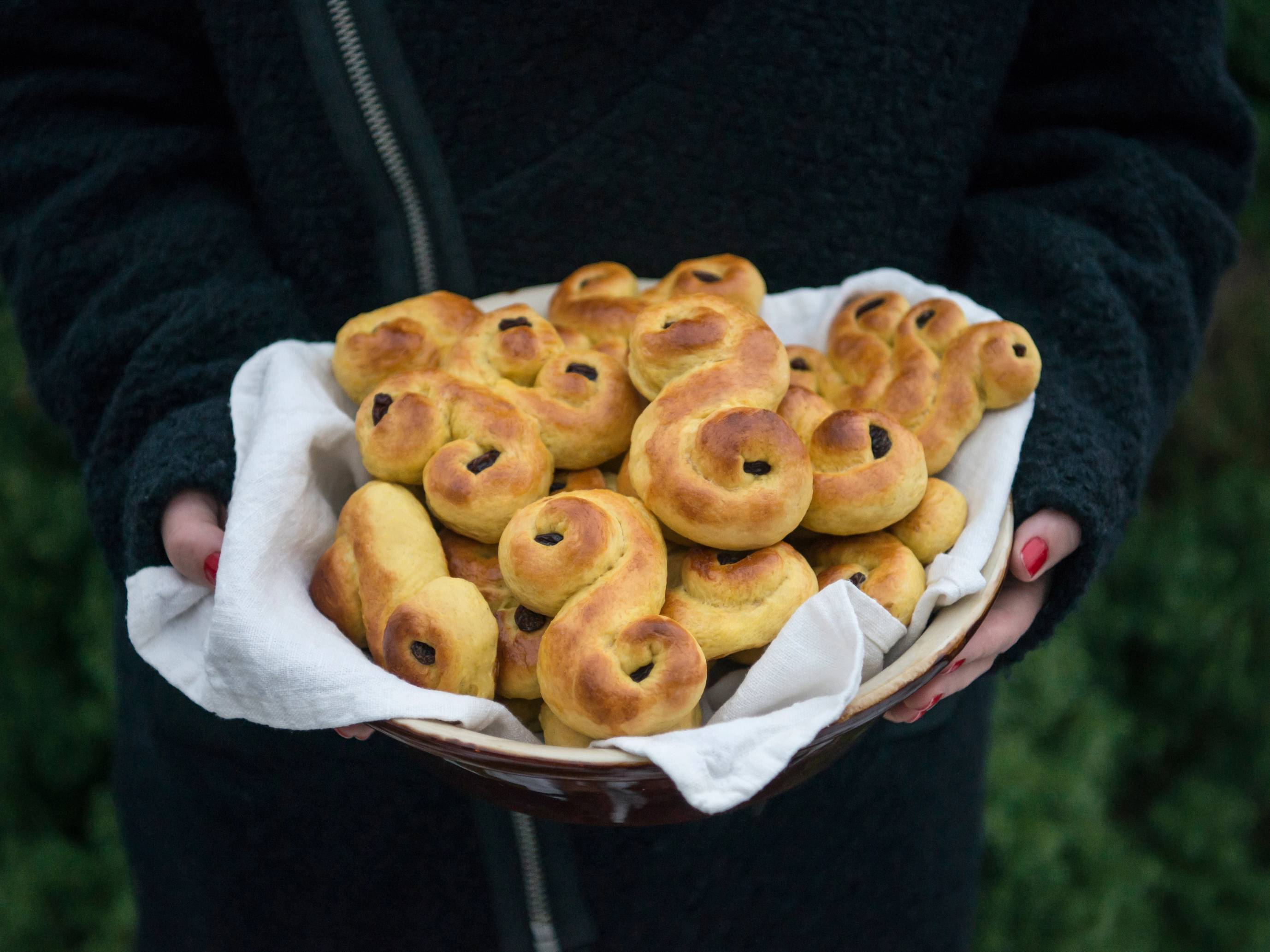 Person holder en tallerken med nybakte lussekatter på, omgitt av grønt gress i bakgrunnen.